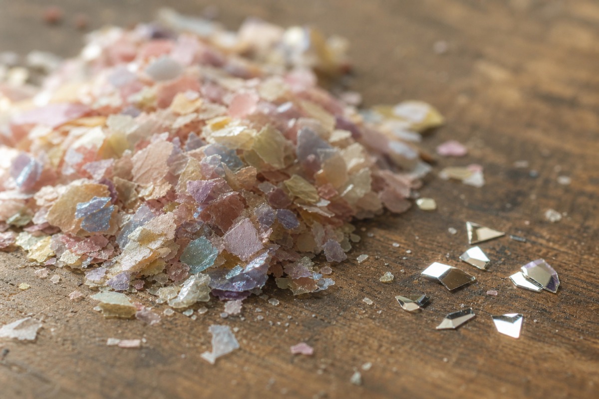 Detalle de un montoncito de bioglitter de celulosa más mate junto a unas cuantas partículas de PET sintético brillantes sobre una superficie de madera.