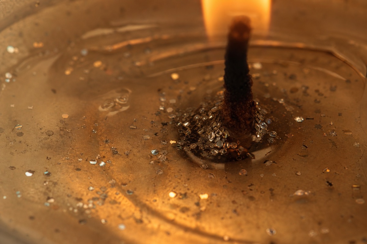 Toma macro de cera transparente fundida junto a la mecha de una vela encendida, en la que descienden diminutas partículas plateadas brillantes.