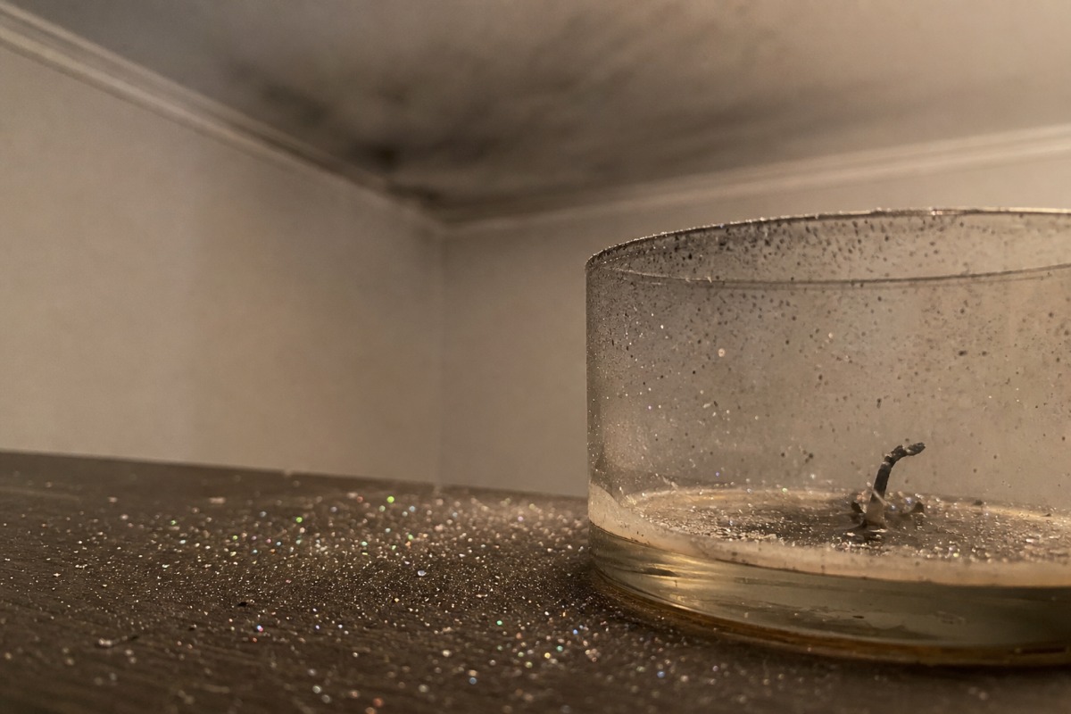 Diminutas partículas brillantes esparcidas por la mesa alrededor de una vela consumida, con una fina película gris de hollín en el techo al fondo.